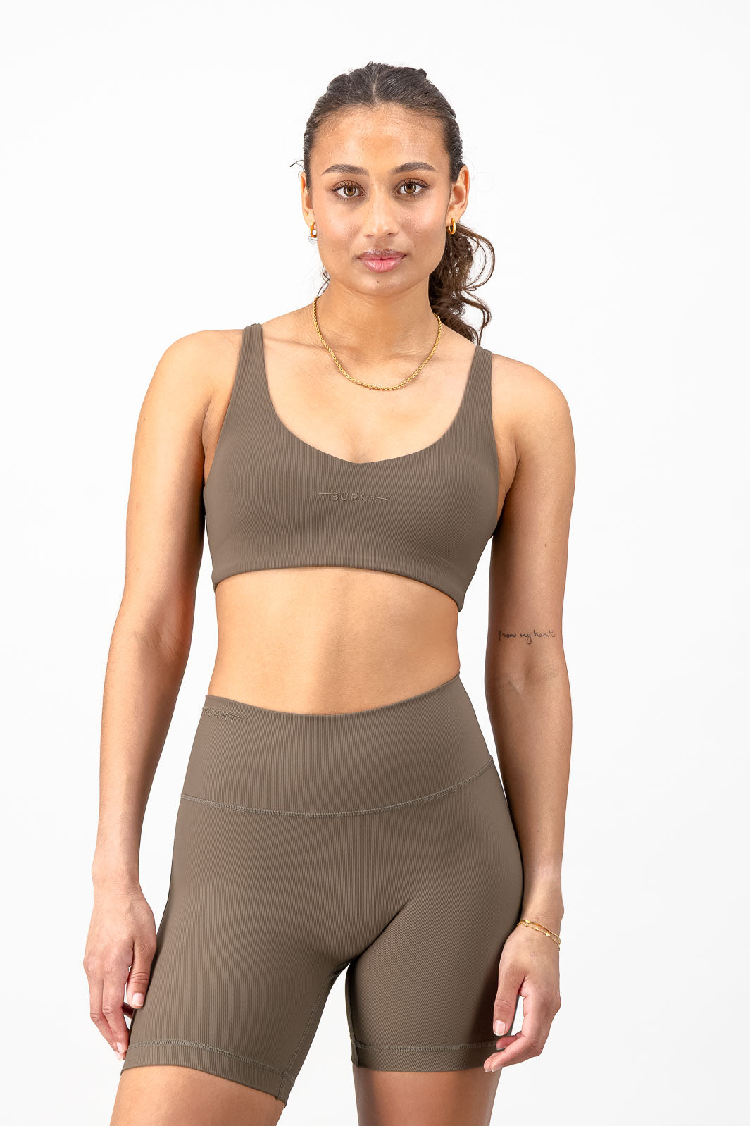 A woman modeling a matching athletic set consisting of a cropped sports bra and high-waisted biker shorts in a solid taupe-brown color, shot against a clean white background.